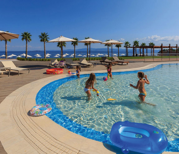 Children playing in a shallow outdoor pool with sea view and palm trees