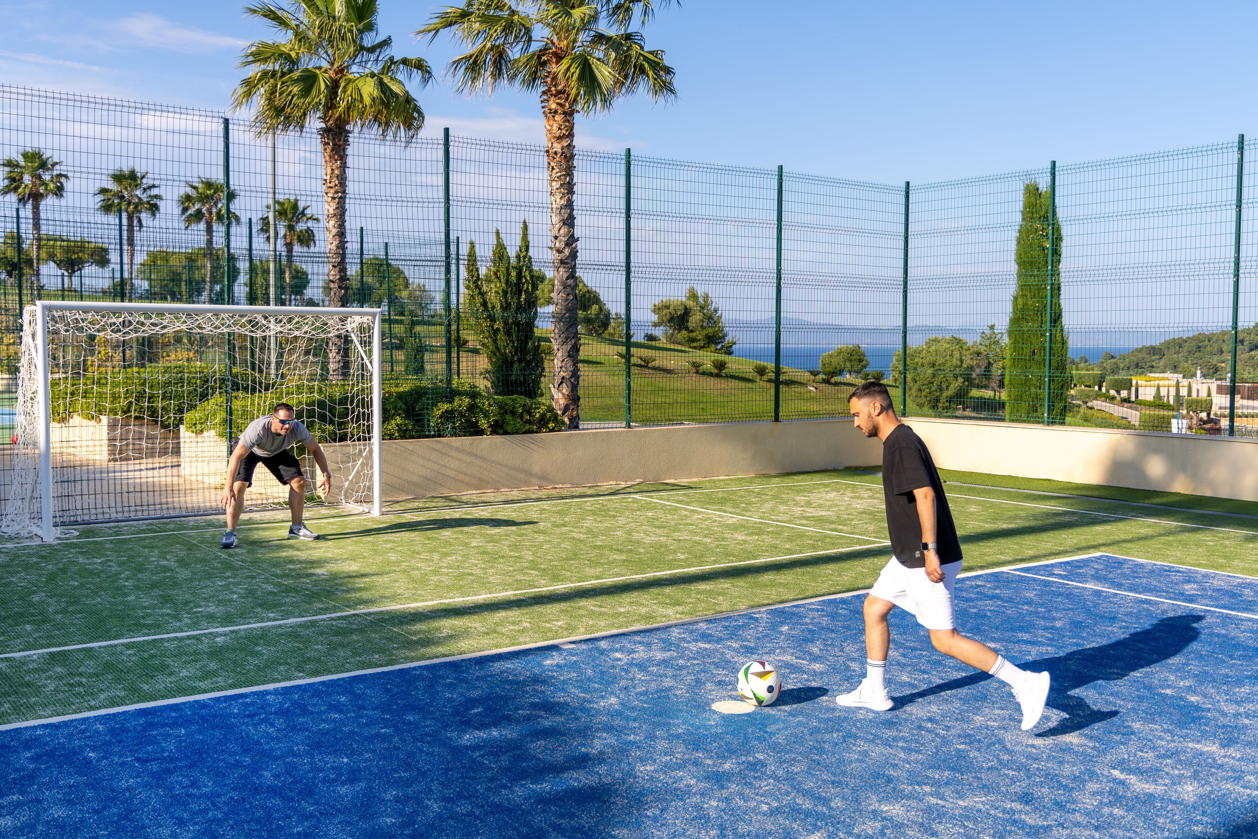 Two men playing football on outdoor court with palm trees and sea view