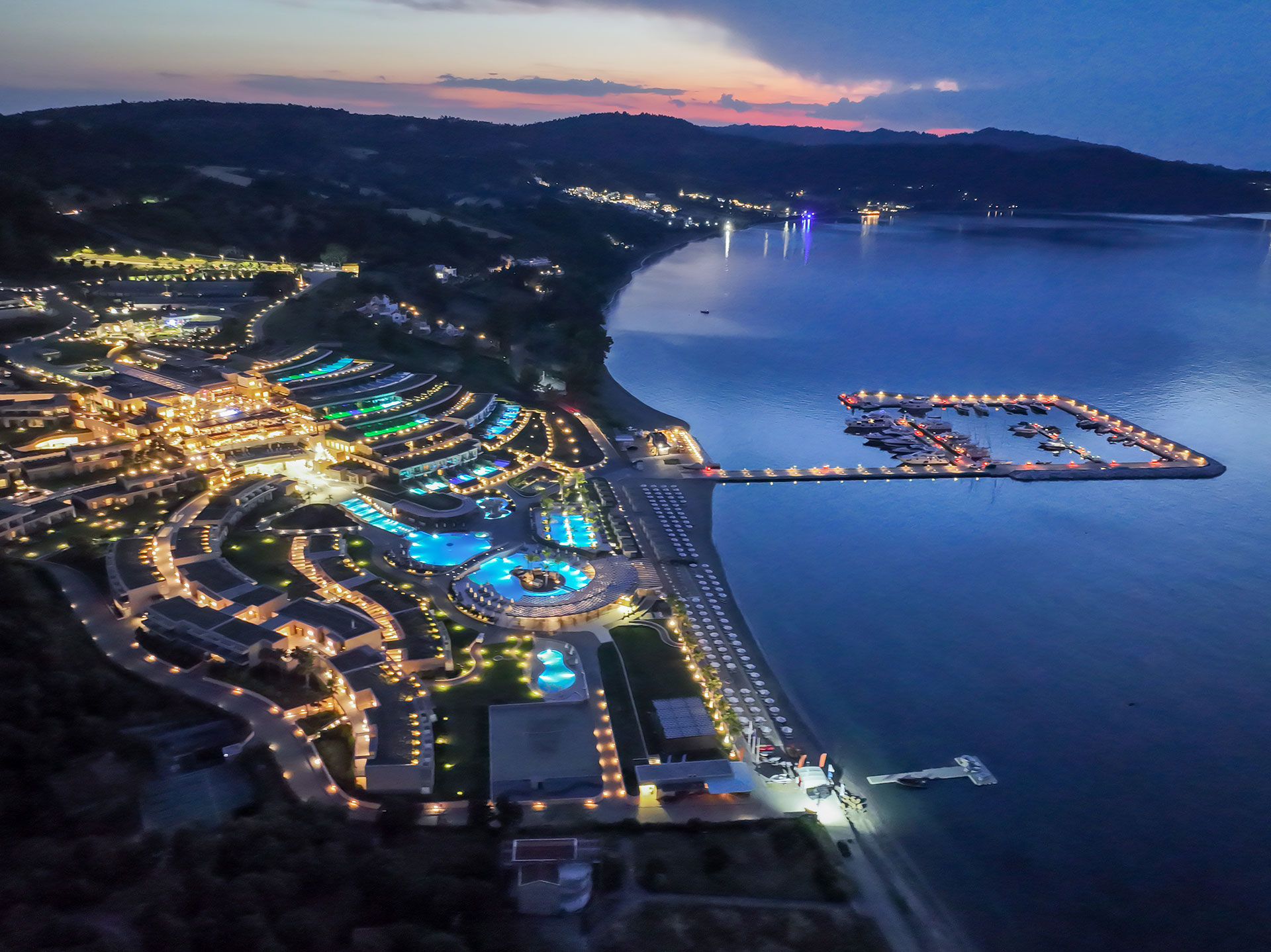 Night aerial view of illuminated pools, marina, and coastline reflecting on calm waters