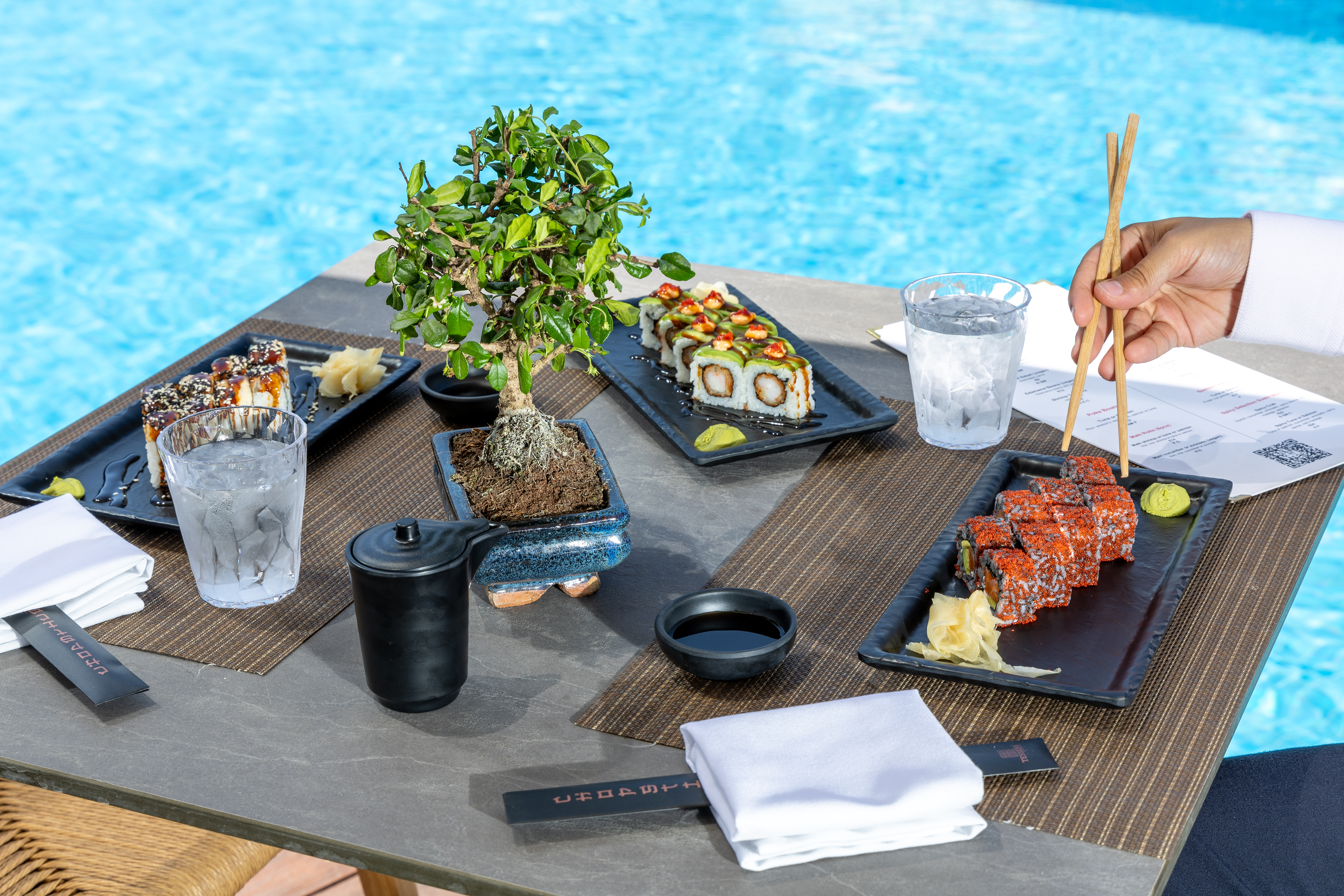 Table with assorted sushi rolls, soy sauce, and bonsai centerpiece by the poolside