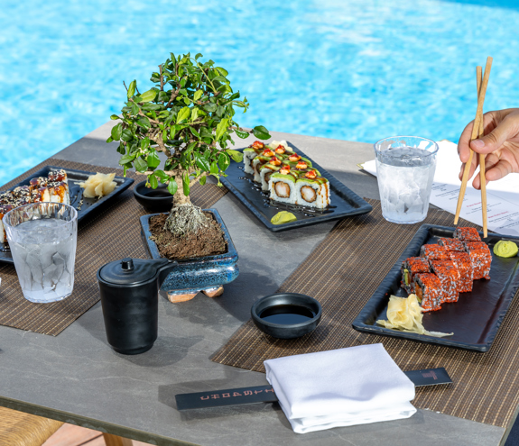Table with assorted sushi rolls, soy sauce, and bonsai centerpiece by the poolside