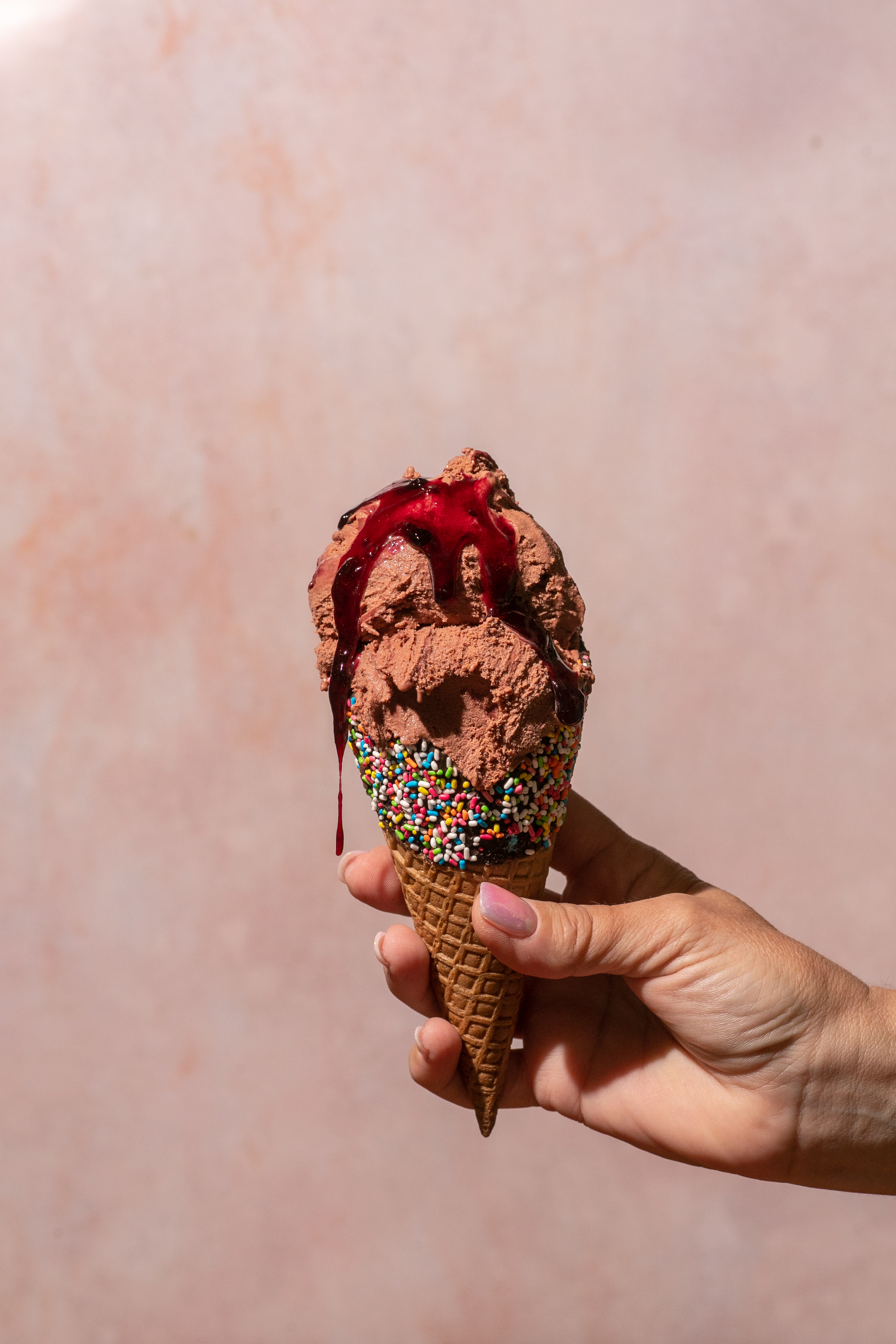 Hand holding Fregio chocolate ice cream cone with berry sauce and rainbow sprinkles