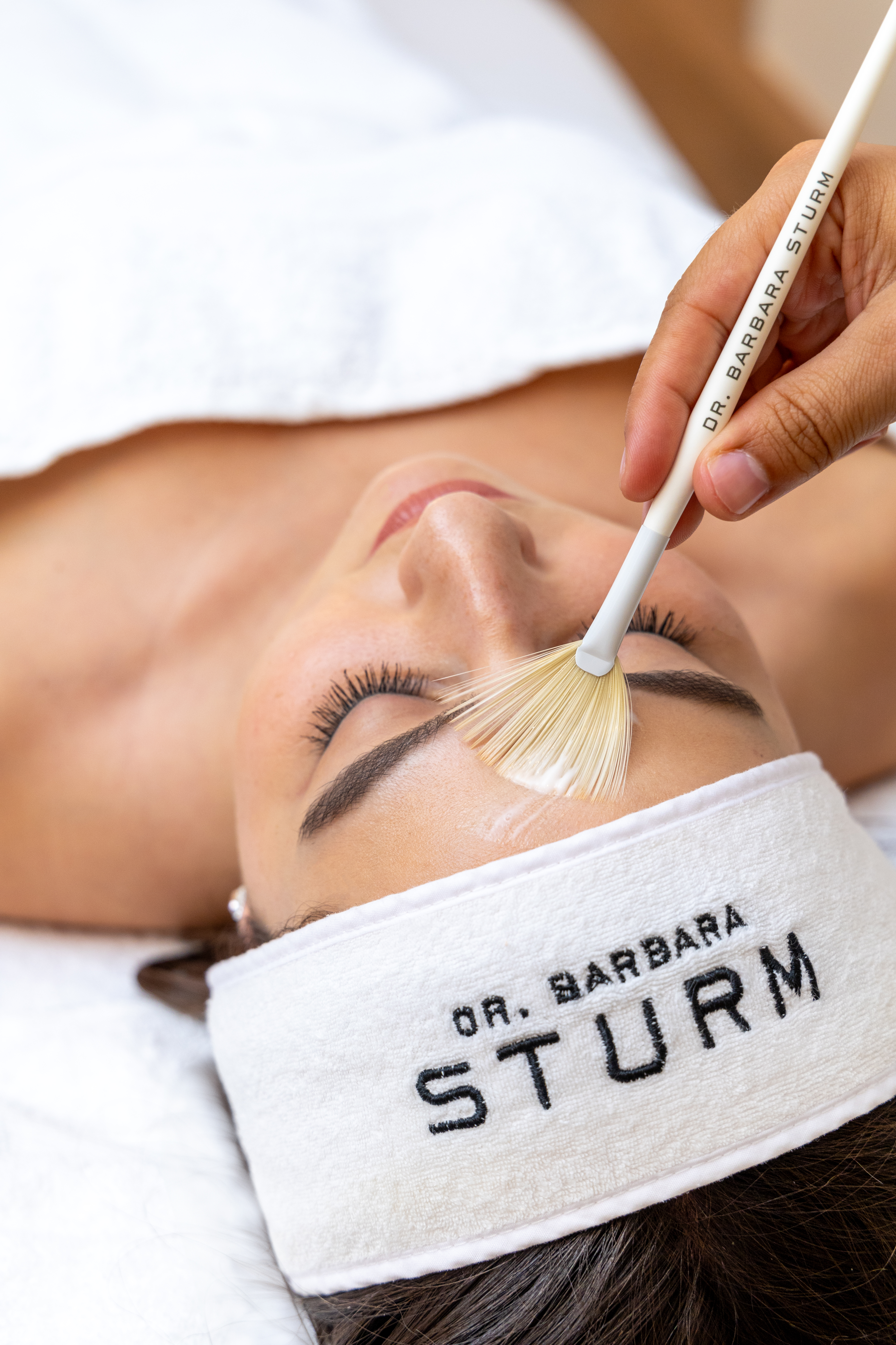 Woman relaxes in spa treatment room while therapist applying skincare product with a brush to her face