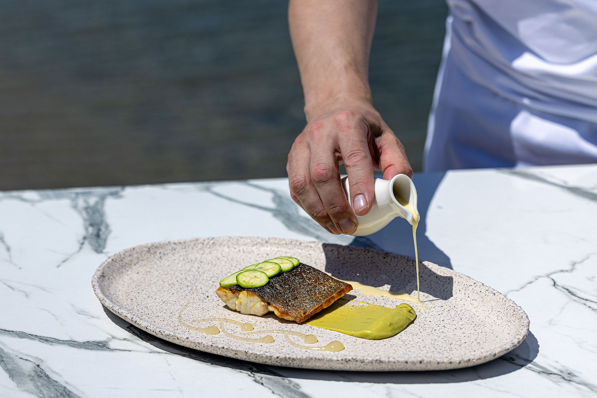 Chef pouring sauce over a grilled fish fillet with cucumber garnish
