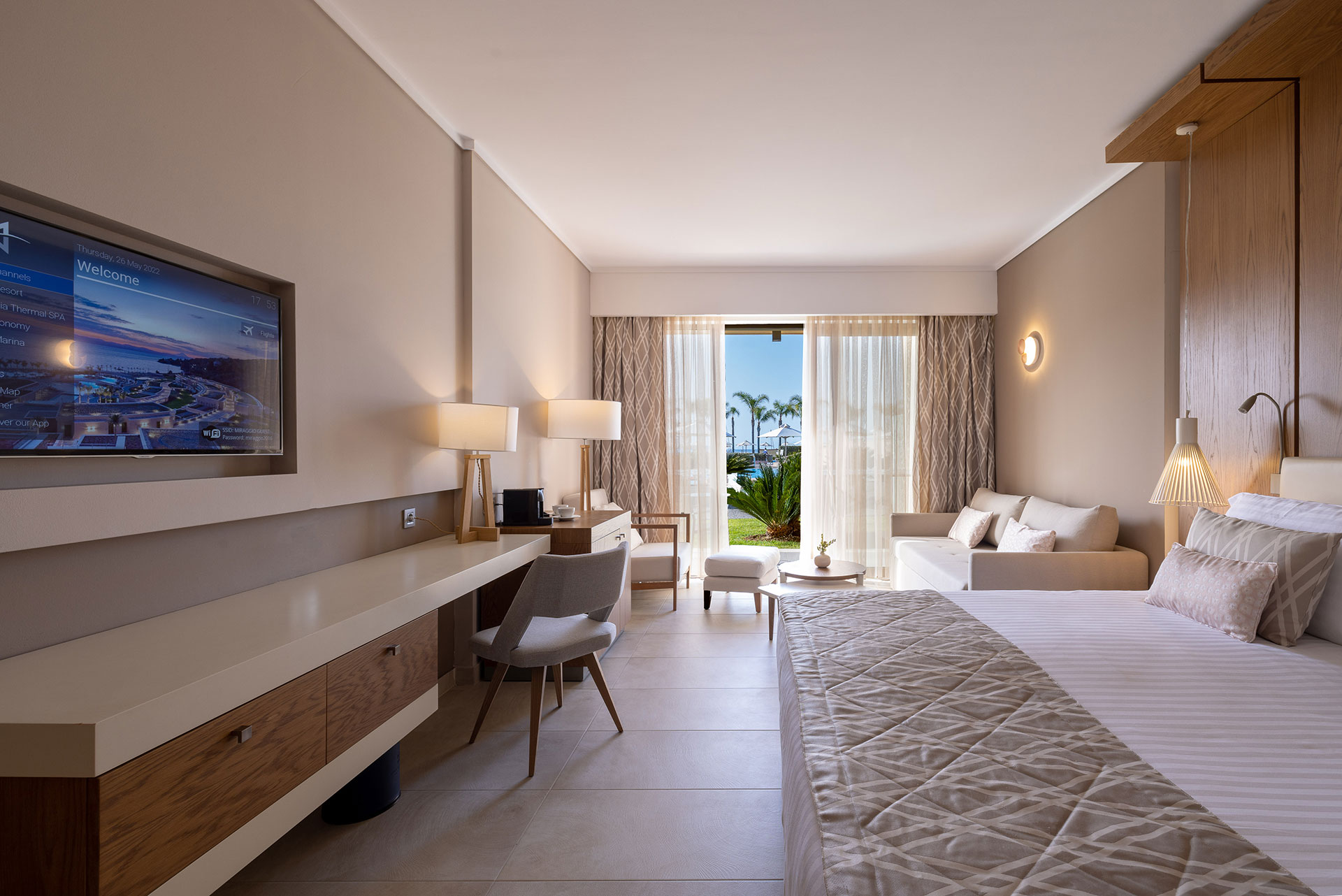 Deluxe room with king size bed, cozy seating area beside the balcony door, and wall mounted TV