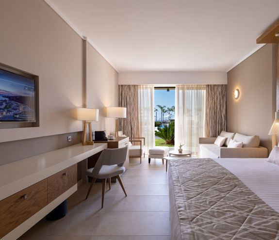 Deluxe room with king size bed, cozy seating area beside the balcony door, and wall mounted TV