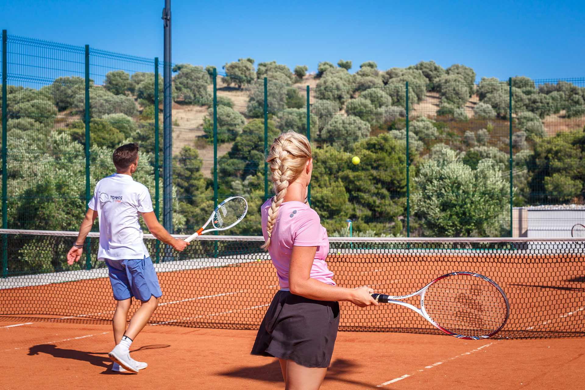 Couple playing tennis on a sunny clay court surrounded by olive trees