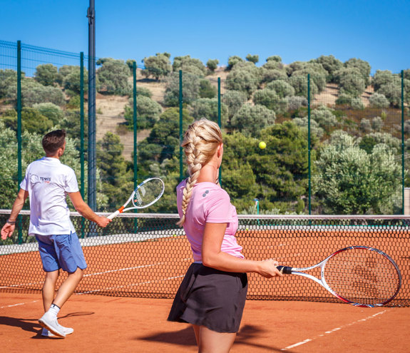 Couple playing tennis on a sunny clay court surrounded by olive trees