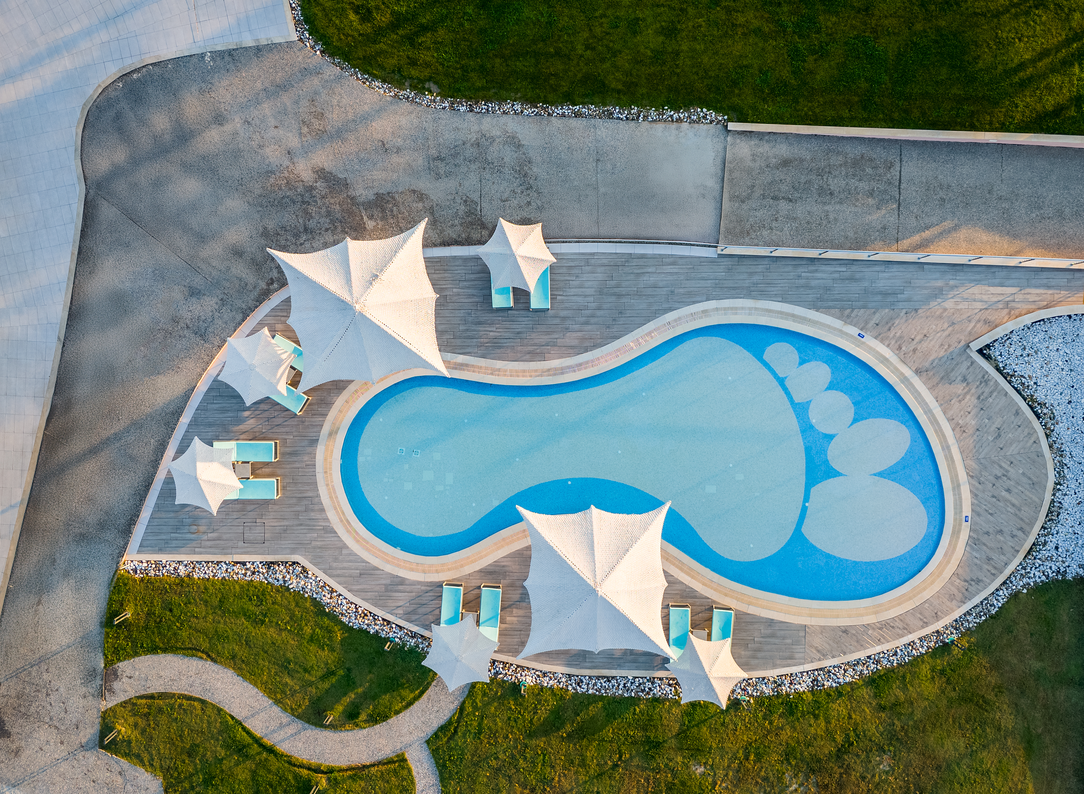 Aerial view of the playful foot-shaped kids' pool with sunbeds and modern white shades surrounded by greenery