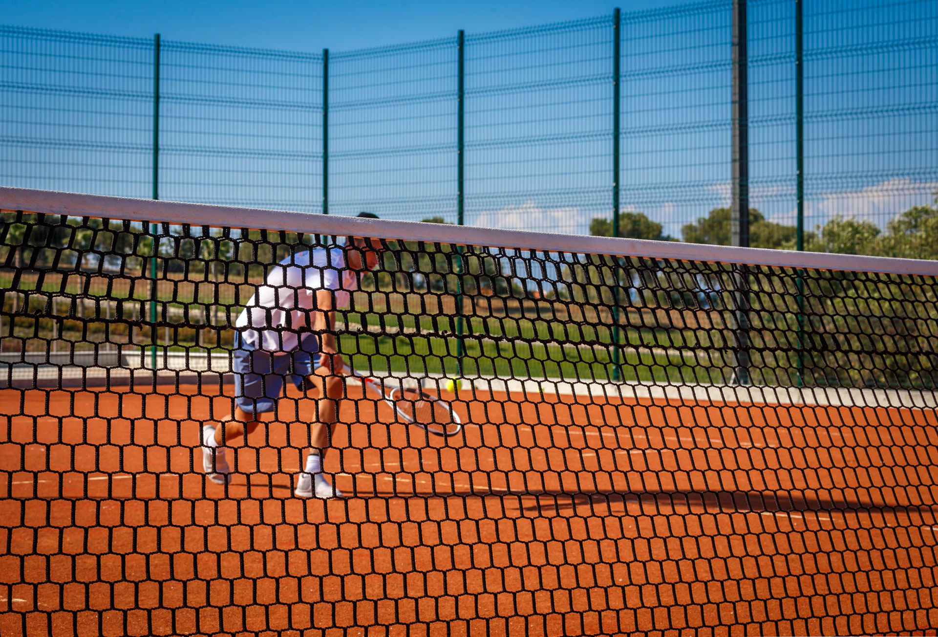 Man playing tennis on a clay court seen through the net on a sunny day