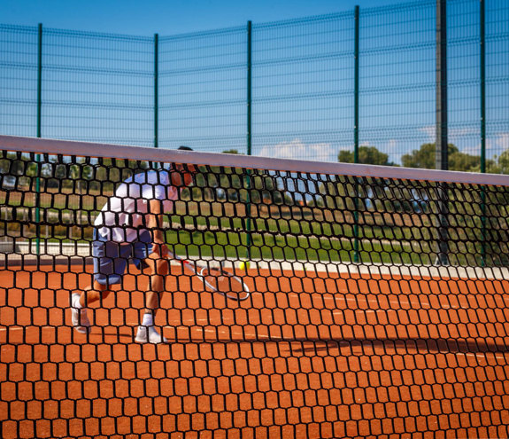 Man playing tennis on a clay court seen through the net on a sunny day