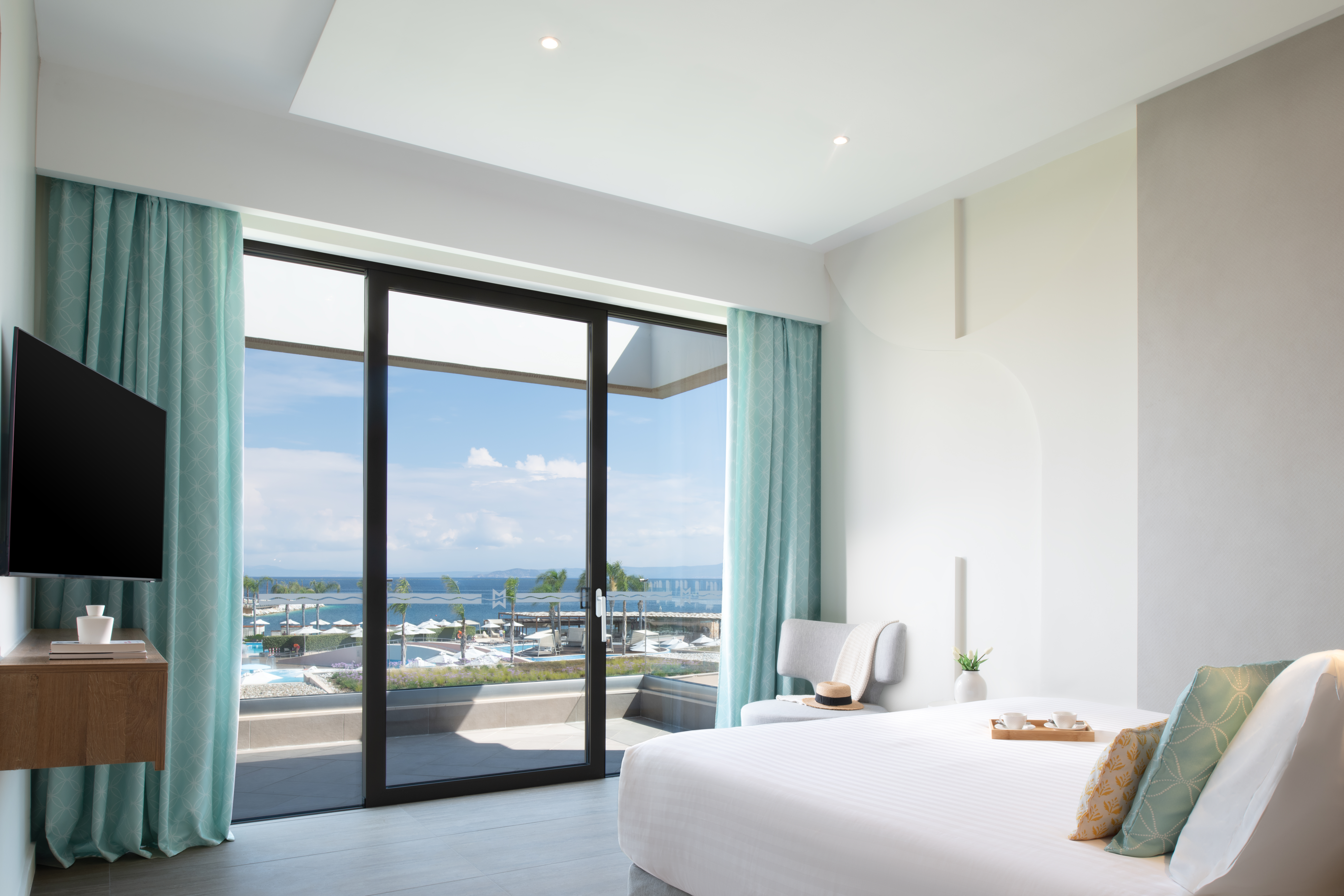 Suite bedroom with king size bed, wall mounted TV, and glass doors overlooking the pools and the sea