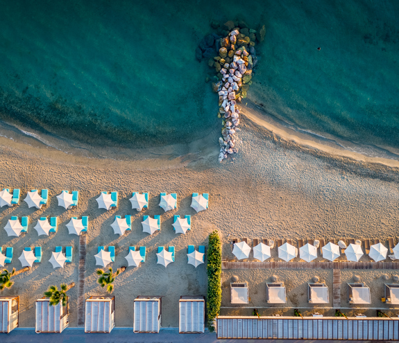Aerial view of the beach with blue sunbeds on the left and bar loungers on the right divided by seaside rocks