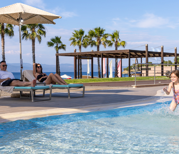 Smiling child splashing in the pool while parents relax on sunbeds with sea views and palm trees