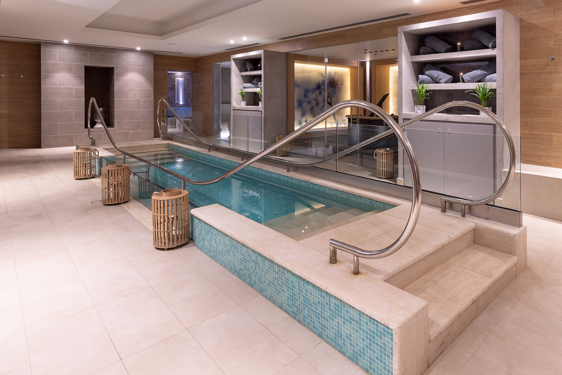 Indoor walking cold-plunge pool with steps and handrails, glass door to sauna and shelves with towels